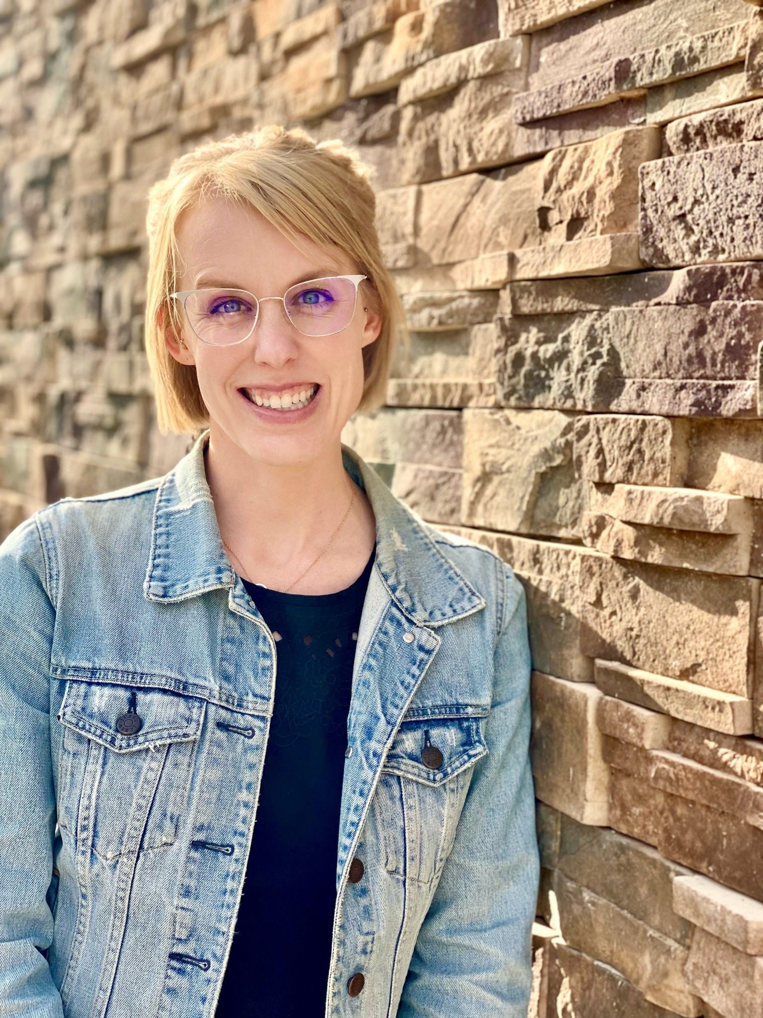 Smiling woman with short blonde hair and glasses stands against a textured stone wall, wearing a denim jacket over a black top.
