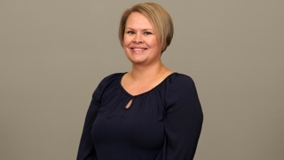 Smiling woman with short blonde hair, wearing a navy blue blouse, poses against a plain beige background. Relevant to team profiles or introductions.